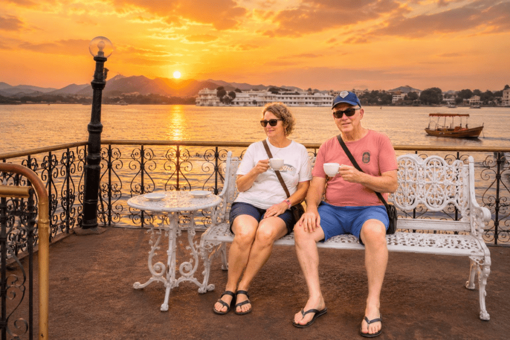 Sarah and Brad sitting together on a bench at sunset on Lake Pichola enjoying a coffee feeling romantic with majestic palaces in the background